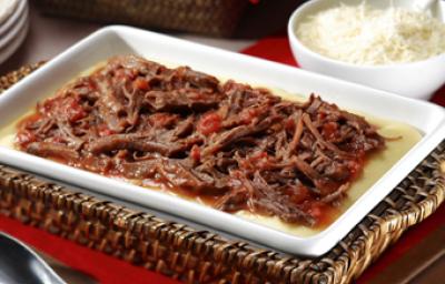 Fotografia em tons de vermelho, branco e marrom, com refratário sobre bandeja de vime com carne desfiada e molho de tomate, sobre guardanapo vermelho, ladeada por pote branco com arroz e uma colher, colher de servir, pratos, tudo sobre bancada de madeira. Fotografia em tons de vermelho, branco e marrom, com refratário sobre bandeja de vime com carne desfiada e molho de tomate, sobre guardanapo vermelho, ladeada por pote branco com arroz e uma colher, colher de servir, pratos, tudo sobre bancada de madeira.