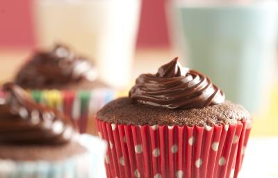 Fotografia em tons de branco e vermelho de uma bancada branca com bolinhas coloridas, sobre ela três cupcakes de Nescau. Ao fundo dois copos com canudos coloridos. Fotografia em tons de branco e vermelho de uma bancada branca com bolinhas coloridas, sobre ela três cupcakes de Nescau. Ao fundo dois copos com canudos coloridos.
