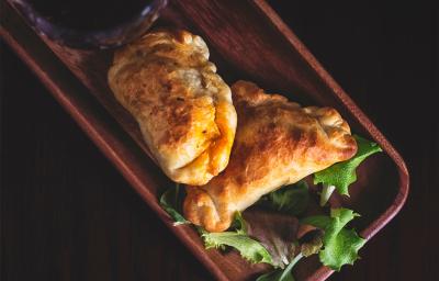 Fotografia em tons escuros vista de cima de duas empanadas recheadas com sardinha sobrepostas e apoiadas em uma tábua de madeira retangular escura. Fotografia em tons escuros vista de cima de duas empanadas recheadas com sardinha sobrepostas e apoiadas em uma tábua de madeira retangular escura.