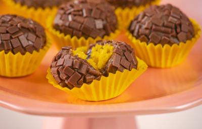 Fotografia em tons de amarelo, rosa e marrom. Ao centro um brigadeiro mordido, mostrando o recheio de bolo de cenoura.