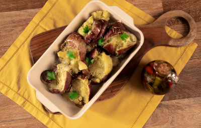 Fotografia em tons de amarelo com uma tábua de madeira ao centro. Em cima da tábua existe uma travessa clara com batatas doces ao murro gratinadas e temperadas com alecrim e azeite Fotografia em tons de amarelo com uma tábua de madeira ao centro. Em cima da tábua existe uma travessa clara com batatas doces ao murro gratinadas e temperadas com alecrim e azeite
