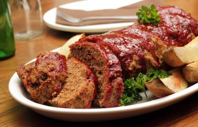 Fotografia de carne com aveia ao forno, metade cortada em fatias e dourada por cima dentro de um refratário branco oval. Ao lado, algumas batatas assadas, e ao fundo, um prato branco com um garfo encima, sobre uma mesa de madeira. Fotografia de carne com aveia ao forno, metade cortada em fatias e dourada por cima dentro de um refratário branco oval. Ao lado, algumas batatas assadas, e ao fundo, um prato branco com um garfo encima, sobre uma mesa de madeira.