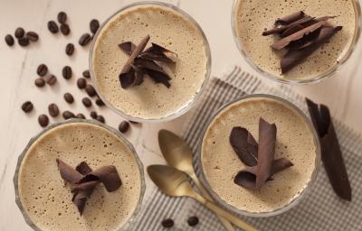Fotografia em tons de branco e marrom de uma bancada branca, sobre ela grãos de café, um paninho branco e preto e quatro taças com mousse de café decorados com chocolate. Fotografia em tons de branco e marrom de uma bancada branca, sobre ela grãos de café, um paninho branco e preto e quatro taças com mousse de café decorados com chocolate.