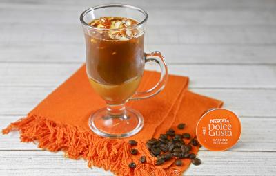 Fotografia em tons de laranja em uma bancada de madeira branca, um pano laranja, uma xícara de vidro com o café com sorvete e pé de moleque dentro dele. Ao lado, uma cápsula de café caseiro intenso Dolce Gusto.