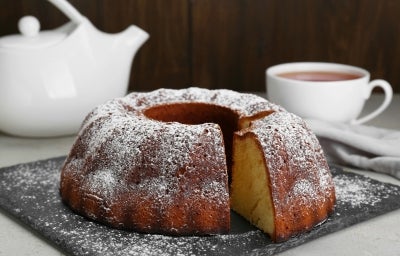 Fotografia de um bolo bundt recém-assado em uma tábua escura, polvilhado com açúcar de confeiteiro. Um bule branco e uma xícara de chá estão ao fundo. Fotografia de um bolo bundt recém-assado em uma tábua escura, polvilhado com açúcar de confeiteiro. Um bule branco e uma xícara de chá estão ao fundo.