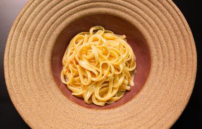 Fotografia em tons de cinza vermelho com um prato vermelho claro ao centro. Dentro do prato existe uma receita de fettuccine Alfredo feita com manteiga e queijo. Fotografia em tons de cinza vermelho com um prato vermelho claro ao centro. Dentro do prato existe uma receita de fettuccine Alfredo feita com manteiga e queijo.