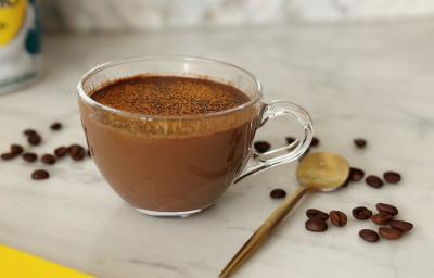 Foto aproximada de uma receita de café cremoso servido em uma xícara de vidro, com uma colher dourada ao lado e alguns grãos de café espalhados na mesa
