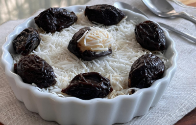 A imagem mostra uma travessa redonda com um creme branco com flocos de coco ralado e ameixas secas pretas decorando. A travessa está apoiada em um guardanapo de pano branco com duas colheres ao fundo.