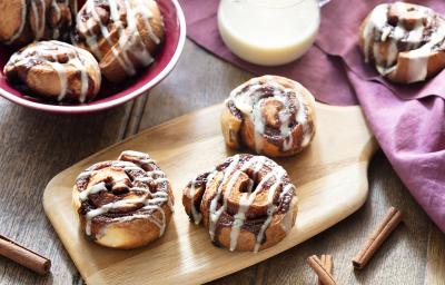 Fotografia em tons de amarelo, em uma bancada de madeira de cor marrom. Ao centro, uma tábua de madeira contendo 3 Cinnamon Rolls. Ao fundo, um pano vermelho e um recipiente contendo cinnamon rolls e uma jarra de leite. Fotografia em tons de amarelo, em uma bancada de madeira de cor marrom. Ao centro, uma tábua de madeira contendo 3 Cinnamon Rolls. Ao fundo, um pano vermelho e um recipiente contendo cinnamon rolls e uma jarra de leite.