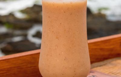 Fotografia de uma taça baixa de vidro com refresco de goiaba com iogurte e NESTON. Dentro da taça tem um canudo de plástico junto com a bebida, que está sobre um apoio de madeira, e ao fundo tem o mar e algumas pedras.