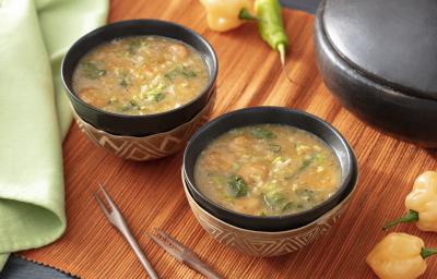 Foto em tons de bege da receita de tacacá servida em duas porções em bowls pretos com adornos em marrom sobre uma esteira de madeira. Ao fundo, há pimentas-de-cheiro e um paninho verde Foto em tons de bege da receita de tacacá servida em duas porções em bowls pretos com adornos em marrom sobre uma esteira de madeira. Ao fundo, há pimentas-de-cheiro e um paninho verde