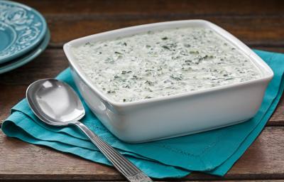 Fotografia em tons de branco e esverdeados de uma bancada de madeira, ao centro um recipiente quadrado está o creme de espinafre em cima de um guardanapo de pano verde água e uma colher de servir ao lado.