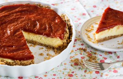 Fotografia em tons de vermelho em uma mesa com uma toalha branca com flores coloridas, um recipiente redondo raso com a cheesecake dentro dele. Ao lado, um prato raso branco com uma fatia da cheesecake. Fotografia em tons de vermelho em uma mesa com uma toalha branca com flores coloridas, um recipiente redondo raso com a cheesecake dentro dele. Ao lado, um prato raso branco com uma fatia da cheesecake.