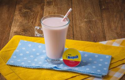 Fotografia em tons de rosa, em uma bancada de madeira de cor marrom. Ao centro, um pano liso amarelo e um pano amarelo quadriculado sobrepostos. Em cima deles, há o frappé de Nesquik com um canudo dentro e ao lado, uma cápsula de Nesquik.