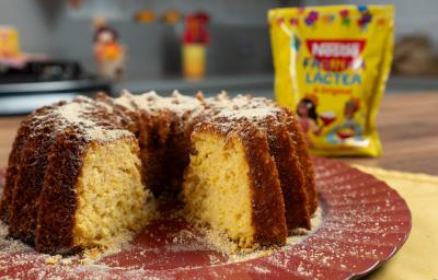 Fotografia de um bolo de milho com farinha láctea apoiado em um prato grande, raso e decorativo de tom vinho. Atrás do bolo tem uma embalagem de farinha láctea. Fotografia de um bolo de milho com farinha láctea apoiado em um prato grande, raso e decorativo de tom vinho. Atrás do bolo tem uma embalagem de farinha láctea.