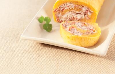Fotografia em tons de amarelo em uma bancada de madeira com um recipiente retangular e o rocambole de mandioca com carne-seca cortado ao meio. Fotografia em tons de amarelo em uma bancada de madeira com um recipiente retangular e o rocambole de mandioca com carne-seca cortado ao meio.