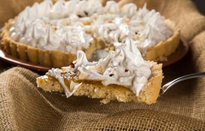 Fotografia de uma espátula segurando uma fatia de torta de cupuaçu e castanha, com merengue por cima. Ao fundo, a mesma torta, inteira sobre apoio redondo na cor marrom. Fotografia de uma espátula segurando uma fatia de torta de cupuaçu e castanha, com merengue por cima. Ao fundo, a mesma torta, inteira sobre apoio redondo na cor marrom.