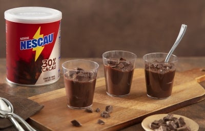 Fotografia de uma tábua retangular de madeira, pequena, com três potinhos de acrílico com o brigadeiro cremoso de colher de nescau. Ao lado tem a embalagem de NESCAU Extra Cacau 30%. Fotografia de uma tábua retangular de madeira, pequena, com três potinhos de acrílico com o brigadeiro cremoso de colher de nescau. Ao lado tem a embalagem de NESCAU Extra Cacau 30%.