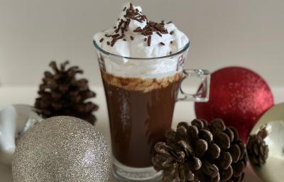 Fotografia de uma xícara de vidro longa com uma receita de espresso cremoso com chocolate, chantilly por cima e um pouco de granulado decorando. A xícara está sobre uma mesa branca, e rodeando da xícara tem alguns enfeites de natal.