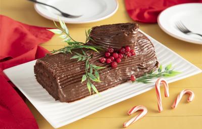 Fotografia em tons de vermelho em uma bancada de madeira amarela, um pano vermelho ao lado e ao centro, um recipiente retangular branco com o rocambole de chocolate em cima com decoração natalina. Fotografia em tons de vermelho em uma bancada de madeira amarela, um pano vermelho ao lado e ao centro, um recipiente retangular branco com o rocambole de chocolate em cima com decoração natalina.