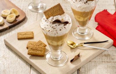 Fotografia em tons de marrom claro com duas taças de vidro ao centro. Dentro de cada taça existe uma receita de banoffee com camadas de biscoito TOSTINES, banana, doce de leite e chantilly. Fotografia em tons de marrom claro com duas taças de vidro ao centro. Dentro de cada taça existe uma receita de banoffee com camadas de biscoito TOSTINES, banana, doce de leite e chantilly.