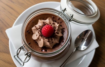 Fotografia de um pote de vidro decorativo com tampa, aberto, com a sobremesa dentro, finalizado com ganache, granulado, chantilly de chocolate e uma framboesa por cima. O pote está sobre um prato de vidro branco, raso, com um paninho branco dobrado. Fotografia de um pote de vidro decorativo com tampa, aberto, com a sobremesa dentro, finalizado com ganache, granulado, chantilly de chocolate e uma framboesa por cima. O pote está sobre um prato de vidro branco, raso, com um paninho branco dobrado.