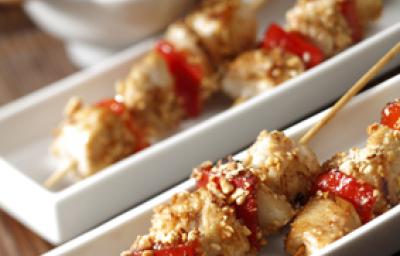 Fotografia em tons de vermelho em uma bancada de madeira com um jogo americano de palha, recipientes brancos retangulares finos com espetinhos de frango com legumes. Ao fundo, um potinho com molho oriental e um paninho vermelho. Fotografia em tons de vermelho em uma bancada de madeira com um jogo americano de palha, recipientes brancos retangulares finos com espetinhos de frango com legumes. Ao fundo, um potinho com molho oriental e um paninho vermelho.