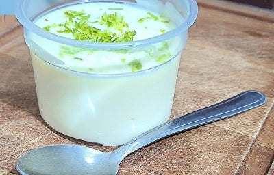 Mousse de limão em pote plástico, finalizada com raspas de limão, sobre tábua de madeira com colher ao lado.