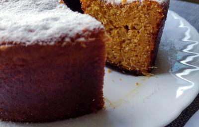 Fotografia em tons de bege em uma bancada com toalha cinza, um prato branco redondo raso, um bolo de milho cortado ao meio com açúcar polvilhado em cima. Fotografia em tons de bege em uma bancada com toalha cinza, um prato branco redondo raso, um bolo de milho cortado ao meio com açúcar polvilhado em cima.
