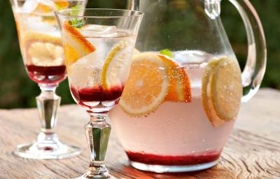 Fotografia em tons de rosa em uma bancada de madeira clara com uma jarra de vidro com o refresco de hibisco e frutas cítricas dentro, decorando com rodelas de laranja. Ao lado, duas taças de vidro com o refresco. Fotografia em tons de rosa em uma bancada de madeira clara com uma jarra de vidro com o refresco de hibisco e frutas cítricas dentro, decorando com rodelas de laranja. Ao lado, duas taças de vidro com o refresco.