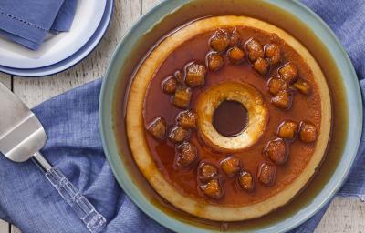 Fotografia em tons de caramelo e azul de uma mesa de madeira vista de cima, ao centro um recipiente redondo azul por cima de um tecido azul, dentro dele está o pudim com a calda de caramelo por cima e decorado com bananas carameladas. Fotografia em tons de caramelo e azul de uma mesa de madeira vista de cima, ao centro um recipiente redondo azul por cima de um tecido azul, dentro dele está o pudim com a calda de caramelo por cima e decorado com bananas carameladas.