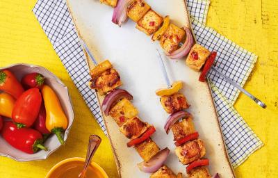 Fotografia em tons de amarelo em uma bancada de madeira amarela, uma tábua de madeira, paninho azul xadrez e três espetinhos de frango. Ao lado, potinhos com pimentas e molho thailandês. Fotografia em tons de amarelo em uma bancada de madeira amarela, uma tábua de madeira, paninho azul xadrez e três espetinhos de frango. Ao lado, potinhos com pimentas e molho thailandês.
