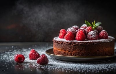 Fotografia de um bolo de chocolate rico, decorado com framboesas vermelhas e mirtilos, polvilhado com açúcar de confeiteiro. Folhas de hortelã adicionam frescor.