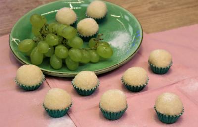 Fotografia em tons de verde em uma bancada de madeira de cor marrom. Ao centro, um pano rosa contendo um prato verde em cima com os brigadeiros e um cacho de uva. Ao redor, há alguns brigadeiros espalhados.