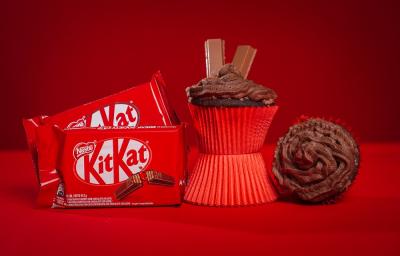 Fotografia em tons de vermelho em uma bancada de madeira de cor vermelha. Ao centro, um cupcake em pé em cima de uma forminha. AO lado direito, um cupcake deitado e ao lado esquerdo, 2 embalagens de kitkat ao laite. Fotografia em tons de vermelho em uma bancada de madeira de cor vermelha. Ao centro, um cupcake em pé em cima de uma forminha. AO lado direito, um cupcake deitado e ao lado esquerdo, 2 embalagens de kitkat ao laite.