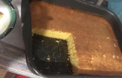 Fotografia de uma forma retangular preta grande com bolo de flocão de milho. Nota-se que faltam quatro pedaços no bolo na forma.