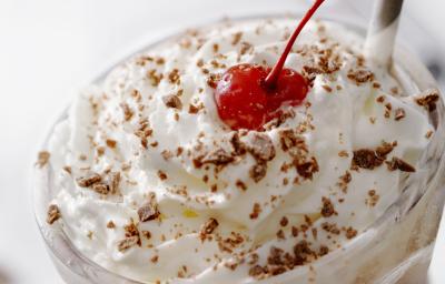 Fotografia de um copo de vidro longo com uma bebida gelada e cremosa de sorvete de chocolate, leite e chantilly. Em cima da bebida, tem raspas de chocolate e um canudo de cores alternadas e uma cereja. Fotografia de um copo de vidro longo com uma bebida gelada e cremosa de sorvete de chocolate, leite e chantilly. Em cima da bebida, tem raspas de chocolate e um canudo de cores alternadas e uma cereja.