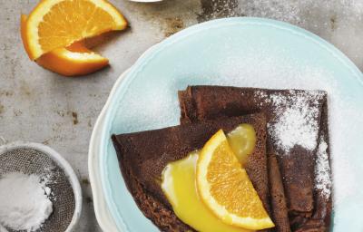 Fotografia de dois crepes de chocolate dobrados recheados com brigadeiro. Por cima, uma calda de laranja por cima, açúcar de confeiteiro polvilhado e meia rodela de laranja. Os crepes estão em um prato azul claro, e ao lado, mais calda de laranja. Fotografia de dois crepes de chocolate dobrados recheados com brigadeiro. Por cima, uma calda de laranja por cima, açúcar de confeiteiro polvilhado e meia rodela de laranja. Os crepes estão em um prato azul claro, e ao lado, mais calda de laranja.