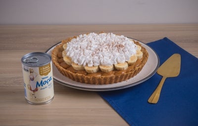 Receita de torta banoffee, servida num prato branco e decorada com chantilly e cacau em pó. A torta está ao lado de uma espátula dourada em uma bancada de madeira com um tecido na cor azul escura Receita de torta banoffee, servida num prato branco e decorada com chantilly e cacau em pó. A torta está ao lado de uma espátula dourada em uma bancada de madeira com um tecido na cor azul escura