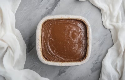 Fotografia vista de cima de um recipiente quadrado com a sobremesa natalina, finalizada com ganache de chocolate sobre uma mesa de mármore com pano branco em volta. Fotografia vista de cima de um recipiente quadrado com a sobremesa natalina, finalizada com ganache de chocolate sobre uma mesa de mármore com pano branco em volta.