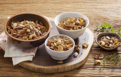 Fotografia em tons de marrom em uma mesa de madeira com uma tábua de madeira redonda, três potinhos com o mix crocante de castanhas temperados com alho e cebola. Fotografia em tons de marrom em uma mesa de madeira com uma tábua de madeira redonda, três potinhos com o mix crocante de castanhas temperados com alho e cebola.