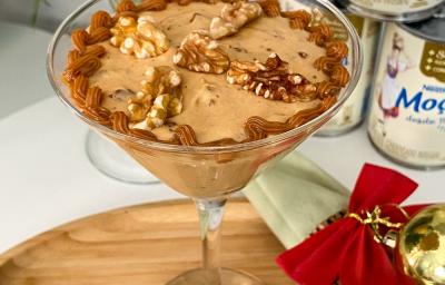 Foto em tons de bege da receita de creme natalino com nozes servida em uma taça de vidro alta sobre uma base de madeira com decoração natalina Foto em tons de bege da receita de creme natalino com nozes servida em uma taça de vidro alta sobre uma base de madeira com decoração natalina