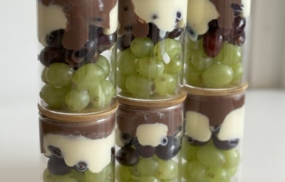 Fotografia em tons claros com vários potes ao centro. Dentro de cada pote existe uma sobremesa feita com uva, creme de leite ninho com iogurte e chocolate meio amargo