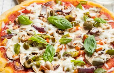 Fotografia de uma pizza com massa de pão sírio com molho de tomate, pedaços de tomate, requeijão e manjericão. A pizza está apoiada sobre uma base de madeira, que está sobre um pano de mesa branco com listras, e ao fundo, dois potinhos e um copo de vidro.
