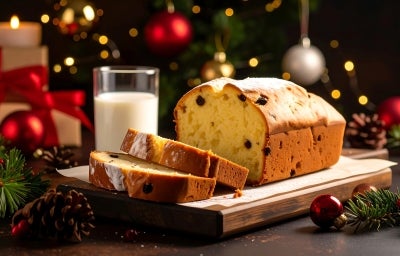 Fotografia de um bolo com frutas secas, parcialmente fatiado, em uma tábua de madeira. O bolo é dourado e fofinho, com passas, é acompanhado por um copo de leite, cercado por decorações natalinas. Fotografia de um bolo com frutas secas, parcialmente fatiado, em uma tábua de madeira. O bolo é dourado e fofinho, com passas, é acompanhado por um copo de leite, cercado por decorações natalinas.