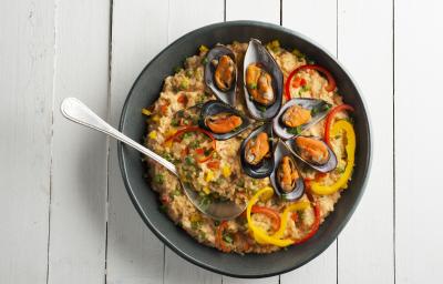 Fotografia em tons de branco e preto de uma bancada branca, sobre ele um prato preto com arroz com frutos do mar e uma colher.