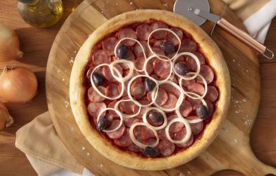 Fotografia em tons de marrom em uma bancada de madeira, uma tábua de madeira redonda com a pizza de calabresa em cima dela. Ao lado, cebola, potinho de azeite e cortador de pizza.