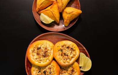 Fotografia em tons de marrom com dois pratos redondos e marrons ao centro. Dentro de um dos pratos existe algumas esfihas fechadas acompanhadas de limão. Dentro de outro prato existe esfihas abertas recheadas de tofu, acompanhadas com limão.