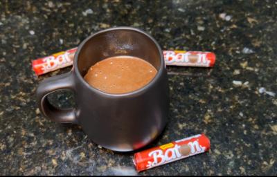 Fotografia em tons de preto com uma caneca preta ao centro. Dentro da caneca existe uma receita de chocolate quente cremosa de cor marrom. Ao lato existe três chocolates em forma de cilindro pequeno da marca BATON Fotografia em tons de preto com uma caneca preta ao centro. Dentro da caneca existe uma receita de chocolate quente cremosa de cor marrom. Ao lato existe três chocolates em forma de cilindro pequeno da marca BATON