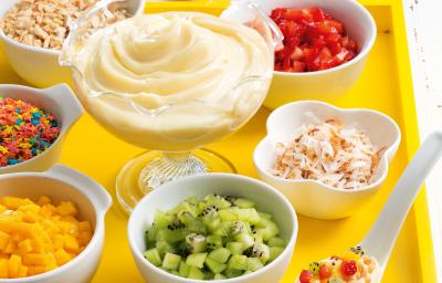 Fotografia em tons de amarelo em uma bancada de madeira clara, uma bandeja amarela com vários potinhos com frutas e doces e ao centro um pote de vidro com o Moça de Colher.